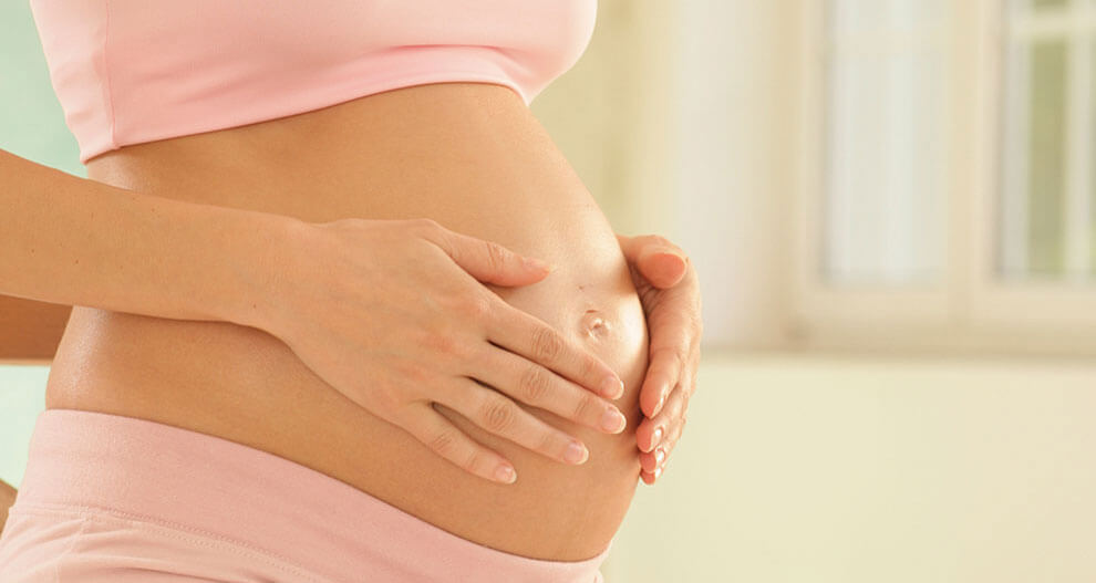 A pregnant woman in pink clothing, sitting by a window, gently cradles her belly.