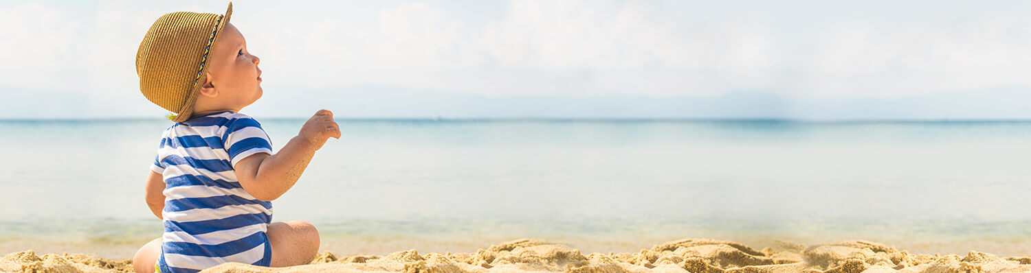 A child in a striped shirt and straw hat sits on a sandy beach, looking out at the ocean.