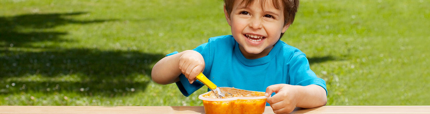 A child in a blue shirt is eating from a yellow container outdoors.