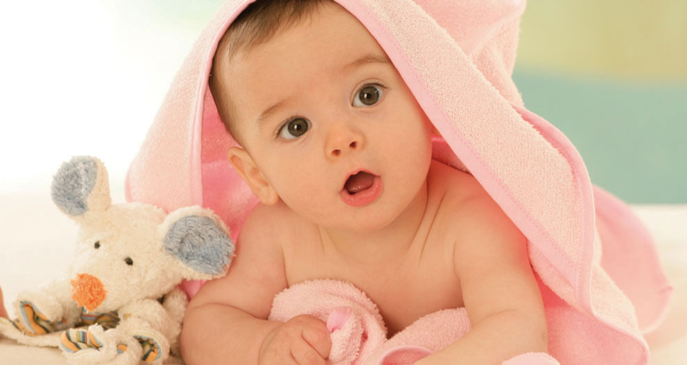 A baby wrapped in a pink towel, lying on a white surface, with a plush toy mouse beside them.
