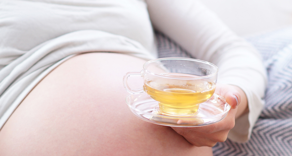 A person is holding a clear glass cup with a yellowish liquid, near their belly.