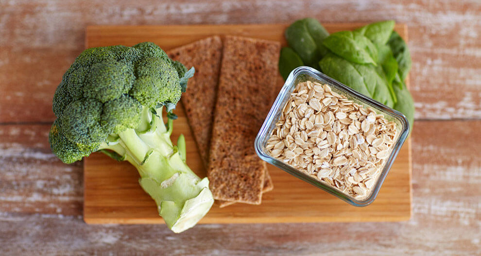 A wooden cutting board holds a stalk of broccoli, a glass container of rolled oats, a pair of brown crackers, and a bunch of fresh green spinach leaves.