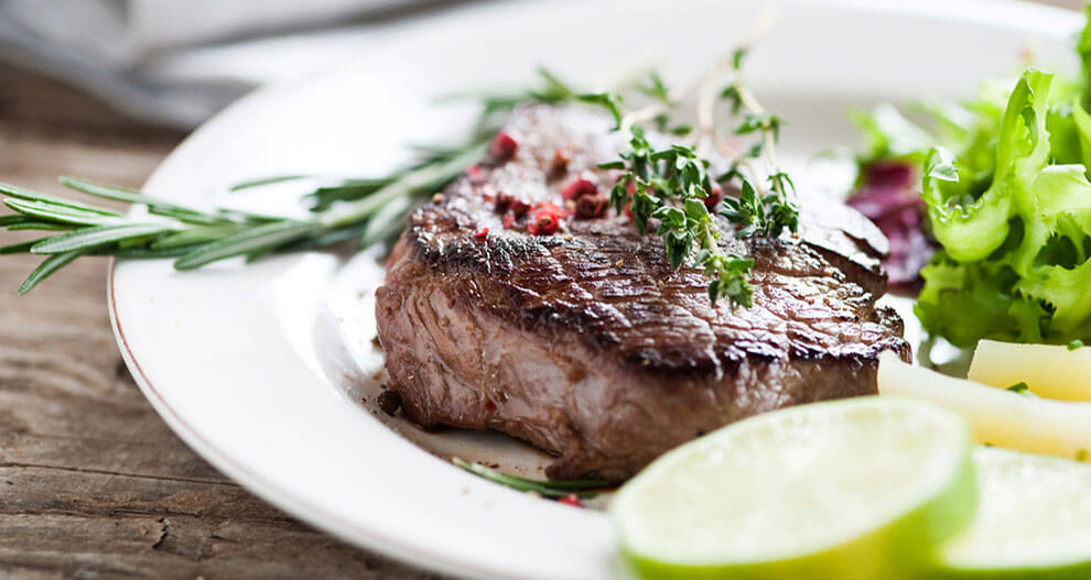 A steak with rosemary and thyme garnish, served alongside a green salad and lime slices.