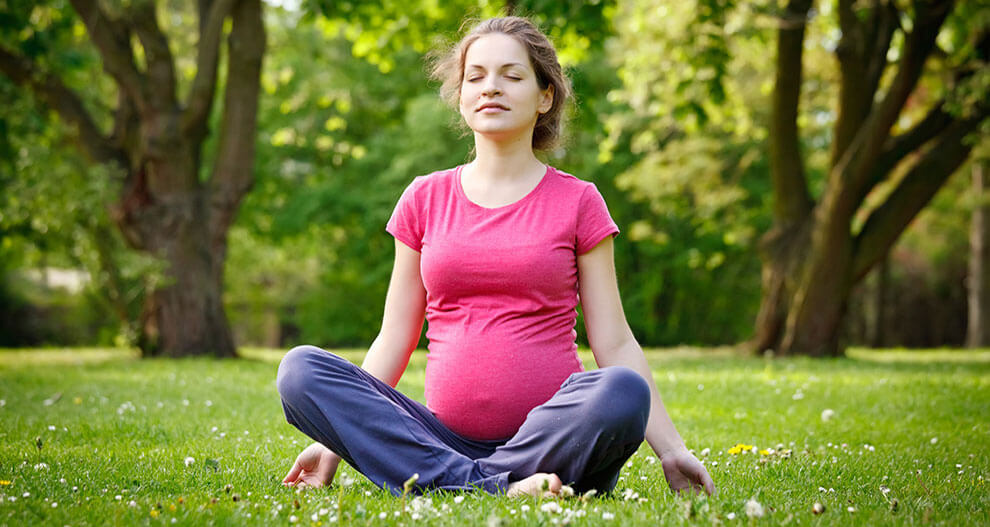 A woman in a pink shirt and blue pants sits cross-legged on grass with flowers, surrounded by trees.