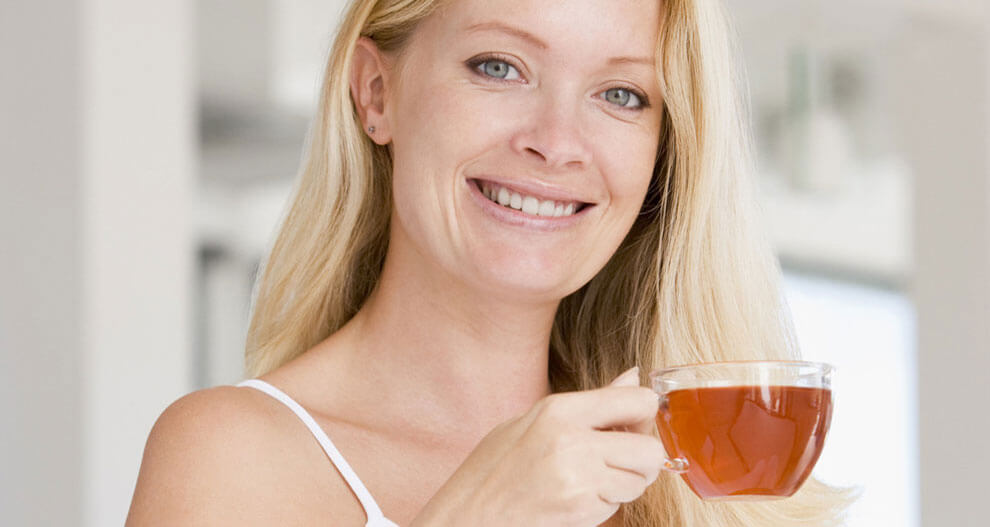 A woman with blonde hair is holding a clear glass cup filled with a reddish-brown liquid, possibly tea or coffee.