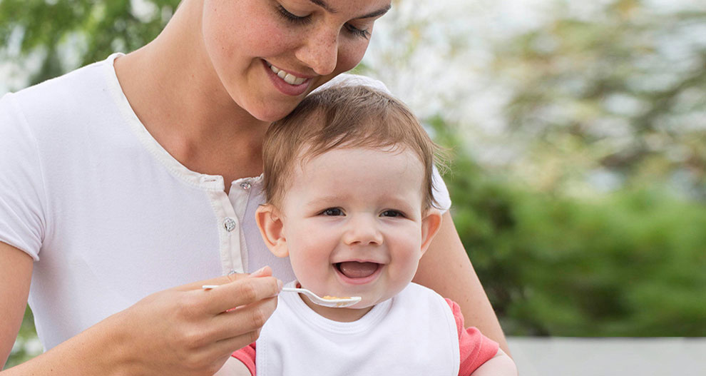 A woman in a white shirt is feeding a baby with a spoon.